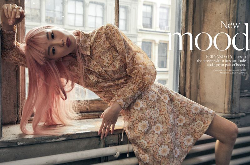 A woman with pink hair leans against a window frame in a floral print dress, with city buildings visible in the background.