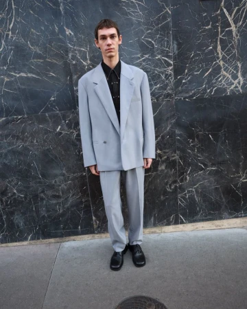 An adult male stands outdoors in a pale grey suit, striking a pose against a dark marble wall on a city street, showcasing men's fashion.