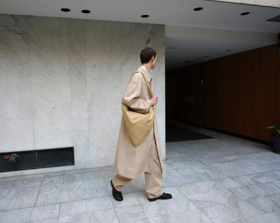 An adult male walks in a beige coat and pants, carrying a beige tote bag, in a building with a marble wall and a dark wood wall.