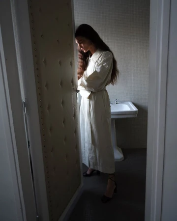 Model in long cream trench coat standing in a tiled bathroom with a pedestal sink for Lemaire SS25 lookbook.