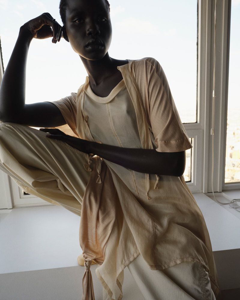 Adult woman with dark skin poses near a bright window, wearing a layered, light-colored cotton outfit; natural daylight.