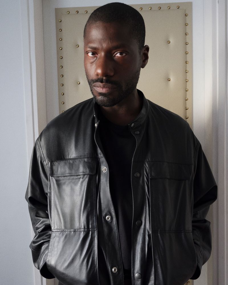 Portrait of a Black man wearing a black leather jacket and dark shirt against a light interior background.
