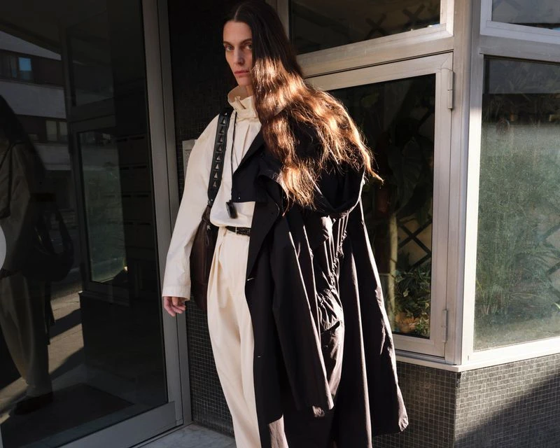 Model with long brown hair wearing a white top and trousers with a black coat for Lemaire SS25