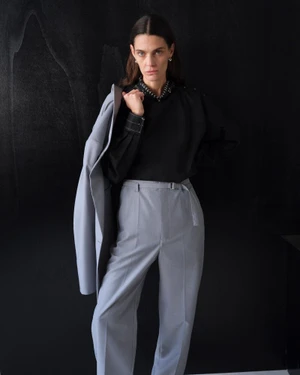 Model wearing a black top and grey trousers holding a light blue blazer against a black textured background for Lemaire SS25