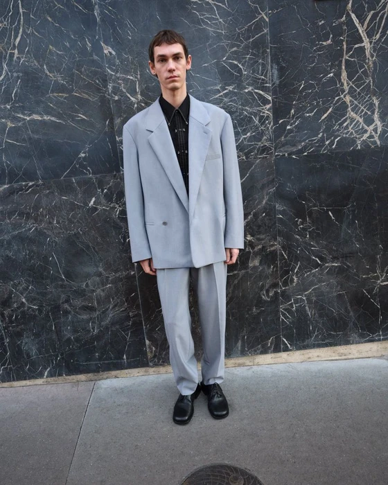 Model standing in a light blue suit and dark shirt against a dark marble wall for Lemaire Lookbook SS25