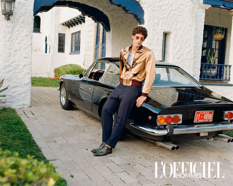 A male model poses with a black vintage car in front of an ornate white building, wearing a gold jacket and sunglasses, L'Officiel Hommes.
