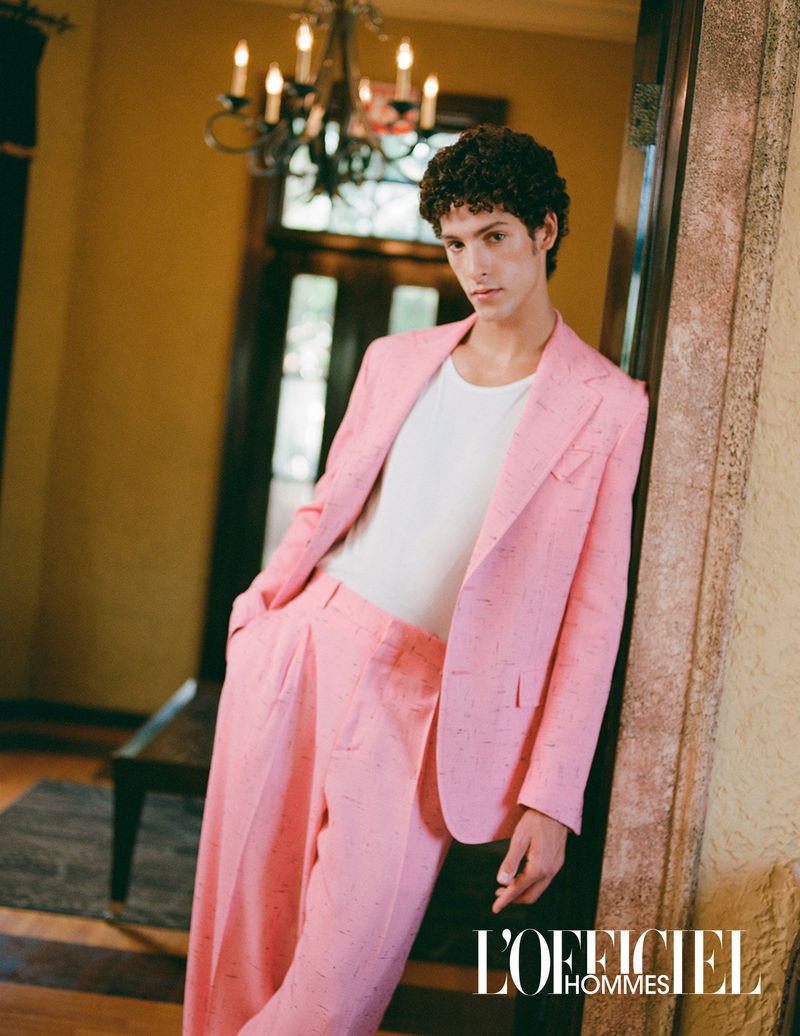 Stylish man in a pink suit poses in an elegant interior. He has curly hair and a white shirt and stands next to a door frame.