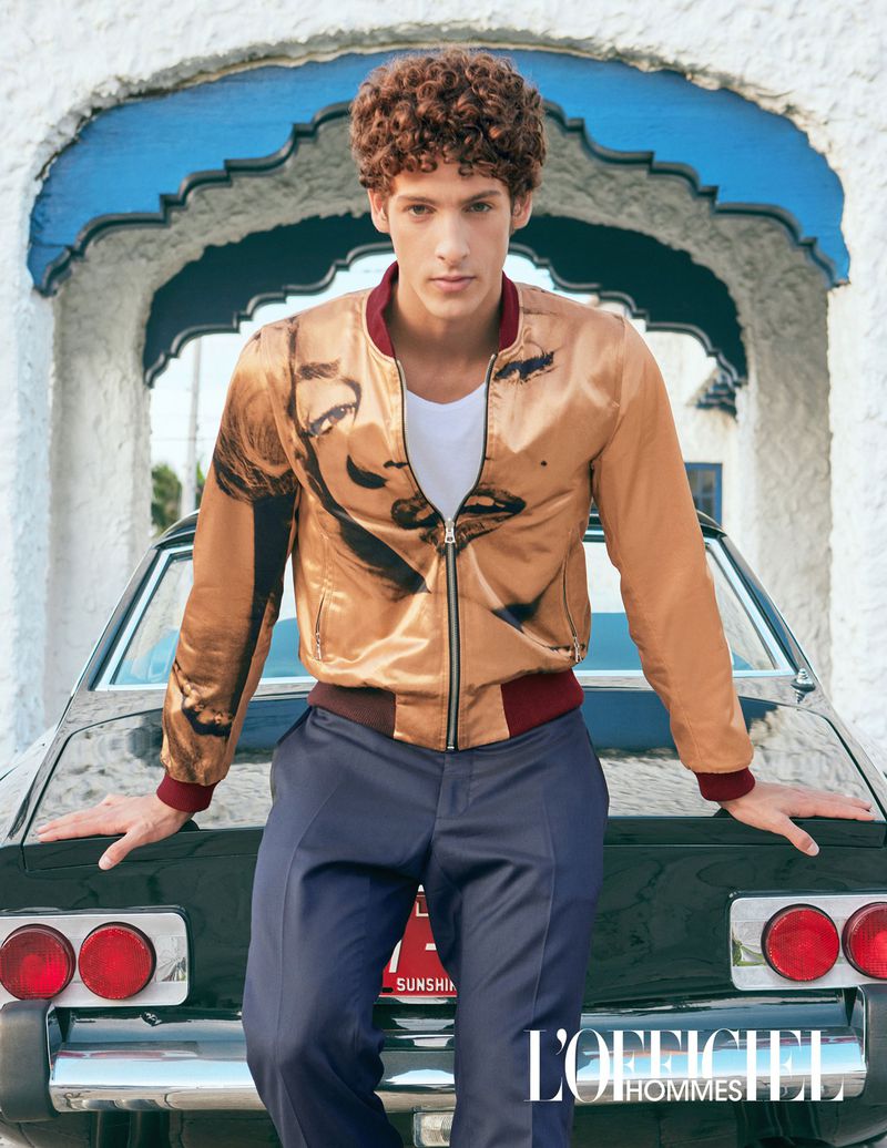 A young man with curly hair poses outdoors in a tan bomber jacket with an image on it, next to a vintage car with a white building archway.