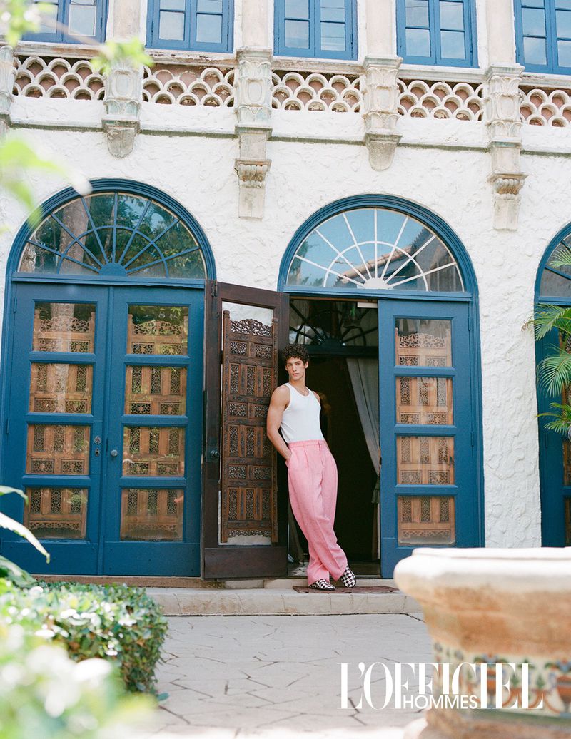 Fashion editorial shot of a young adult man in a white tank top and pink pants leaning against an ornate door, posing for the camera.