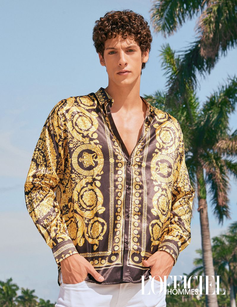 Male fashion model poses outdoors wearing a Versace-patterned silk shirt, white pants, with palm trees and blue sky backdrop.