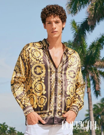 Male fashion model poses outdoors wearing a Versace-patterned silk shirt, white pants, with palm trees and blue sky backdrop.