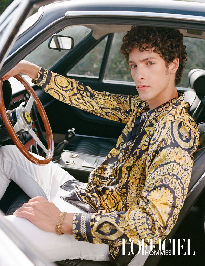 Stylish man with curly hair, wearing a patterned shirt and white pants, sits in a vintage car, creating a fashionable automotive editorial.
