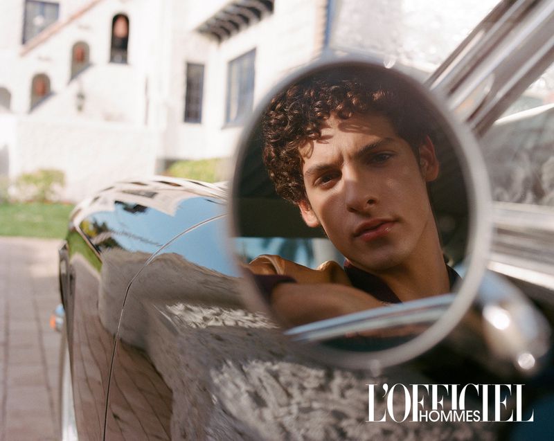Close-up of a young man's face reflected in a car's side mirror with a white stucco building visible in the background on a sunny day.