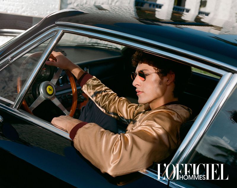 A man in sunglasses sits inside a vintage black Ferrari with a wooden steering wheel, photographed for L'Officiel Hommes magazine.
