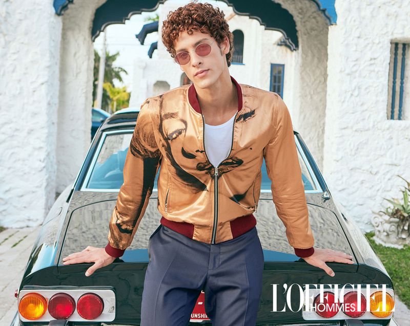 A light-skinned male model with curly hair and sunglasses poses with a vintage car in a fashion editorial shoot, with L'Officiel Hommes logo.