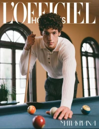 A young man with curly hair in a white knit polo leaning over a pool table with a cue stick and billiard balls.