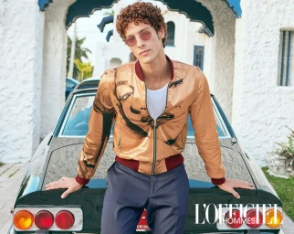 Man with curly hair wearing tinted sunglasses and a gold graphic bomber jacket leaning against a vintage car outside a white villa.