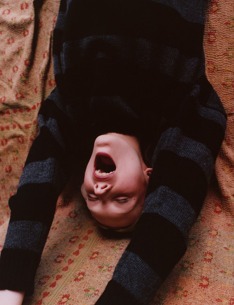 Person with mouth open lies on patterned textile surface wearing striped long-sleeve shirt, captured in eye-level shot indoors.
