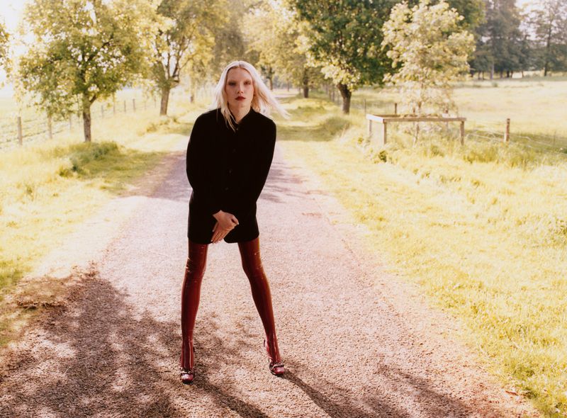 Young woman in a black coat and red thigh-high boots stands in the middle of a gravel road lined with trees in the countryside.