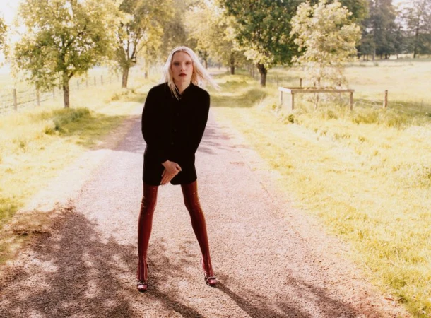 Young woman in a black coat and red thigh-high boots stands in the middle of a gravel road lined with trees in the countryside.