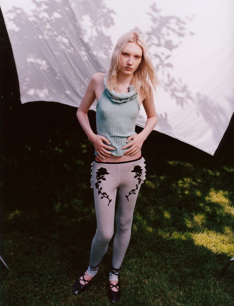 A slender woman with fair skin and light hair poses in a knitted halter top and printed leggings on a lawn with a backdrop.