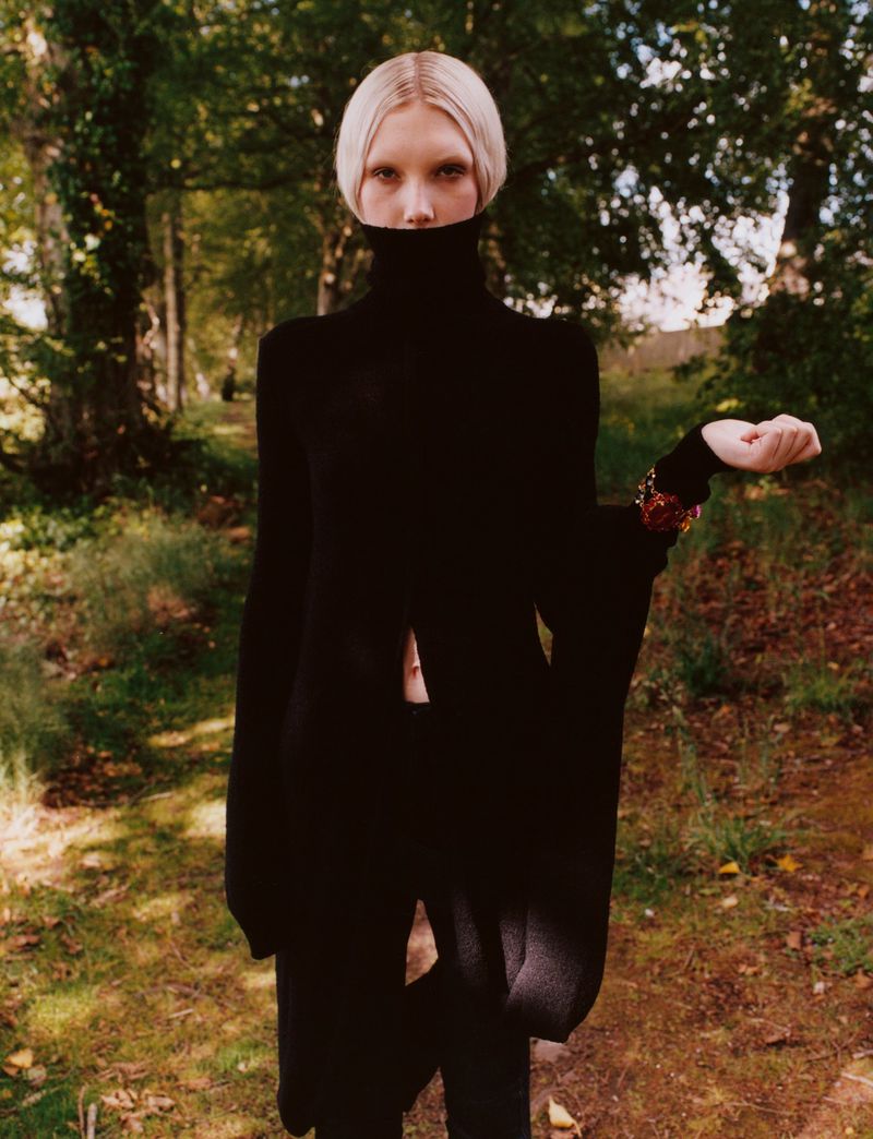 A slender young woman with fair hair stands in a wooded area. She is wearing a long black high-necked dress and bracelet.