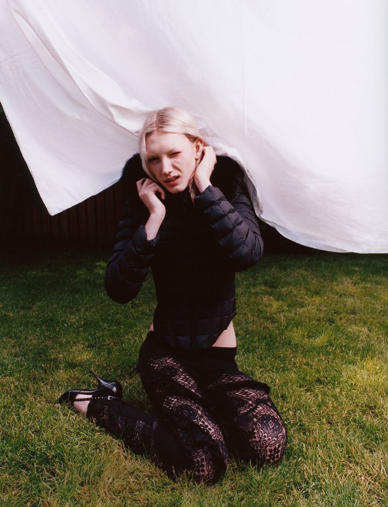 Woman with light blonde hair wearing a black puffer jacket and lace pants kneeling on grass in an outdoor setting with a white sheet backdrop.