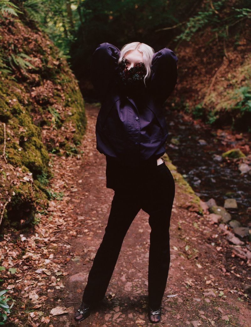 Woman in black clothes and floral face covering stands on a path in a forest, with dappled sunlight and moss-covered ground.