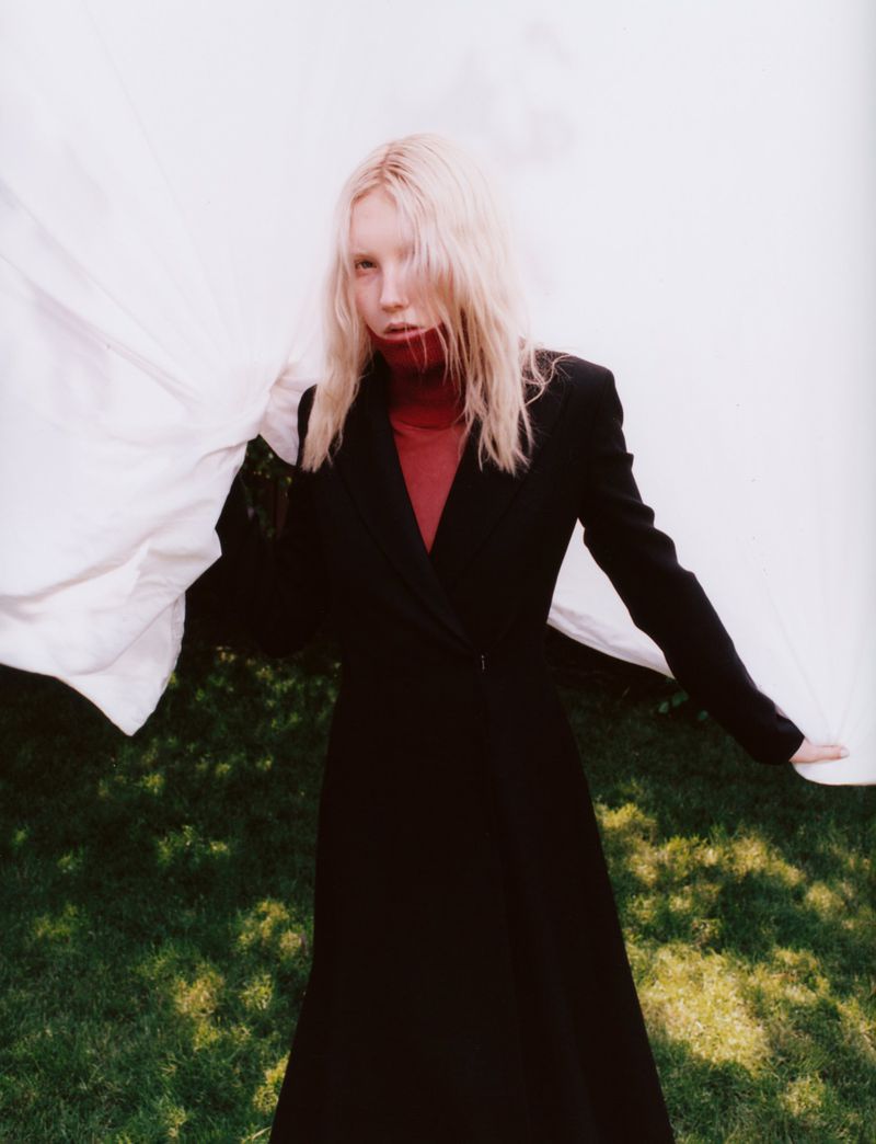 A model with wet blonde hair in a long black coat stands on a grassy lawn with a white cloth draped behind her, looking at the camera.