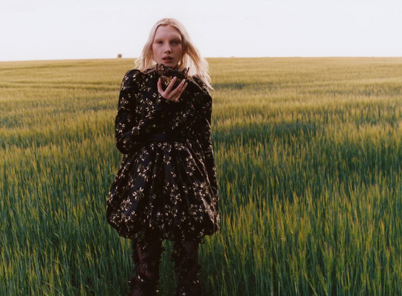 Young woman in a black floral dress stands in a green field holding dried flowers, portrait style, outdoors fashion editorial.