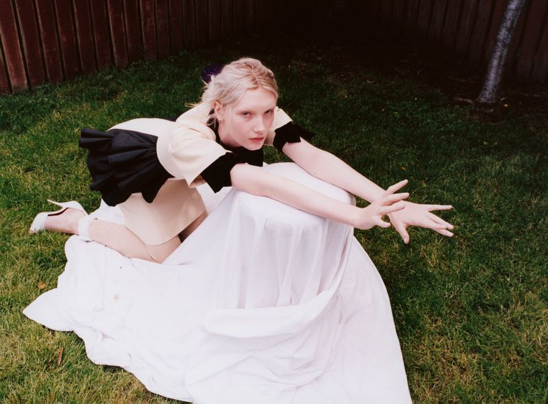 An adult female poses outdoors on a white sheet in a short beige dress with dark accents and white heels against a green lawn and wooden fence.