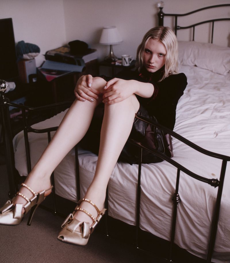 A young adult female with blonde hair is sitting on a bed indoors with golden shoes, wearing a dark top and skirt, with a somber expression.