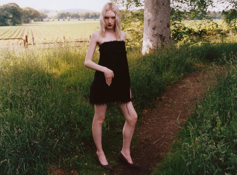 A blonde-haired woman in a short black dress and heels poses on a grassy path in a field on a sunny day. Fashion editorial.