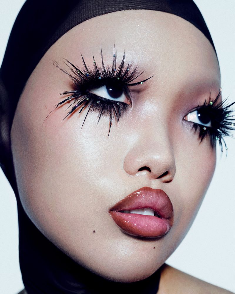 Close-up studio shot of a woman with dramatic eyelashes, glossy lips, wearing a black head covering against a bright background.