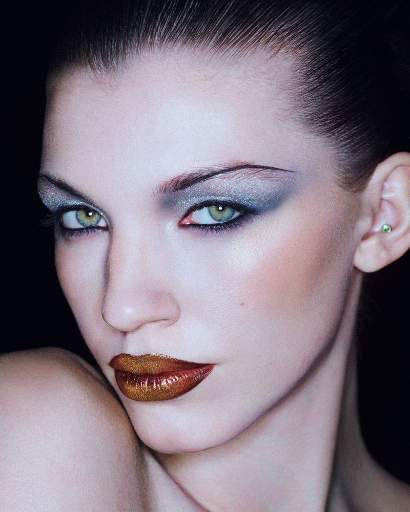 Close-up portrait of a woman with green eyes, metallic gold lipstick, and blue eyeshadow in a dark studio setting for a beauty campaign.