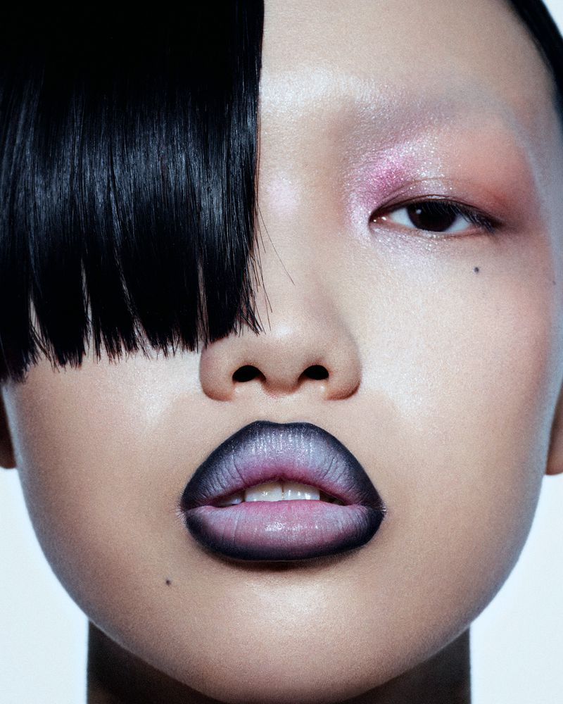 Close-up beauty shot of an Asian female model with ombre lipstick, black hair, and shimmering pink and peach eye shadow, set against white backdrop.