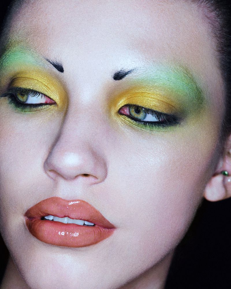 Close-up shot of a female model showcasing vivid green and gold eyeshadow, glossy peach lips, and striking makeup in a studio setting.