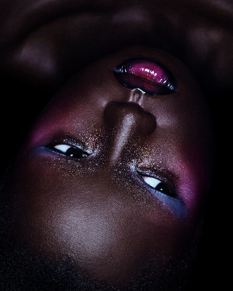 Close-up portrait of a woman with dark skin and glittery makeup on her face, lips and eyes, set against a dark background.