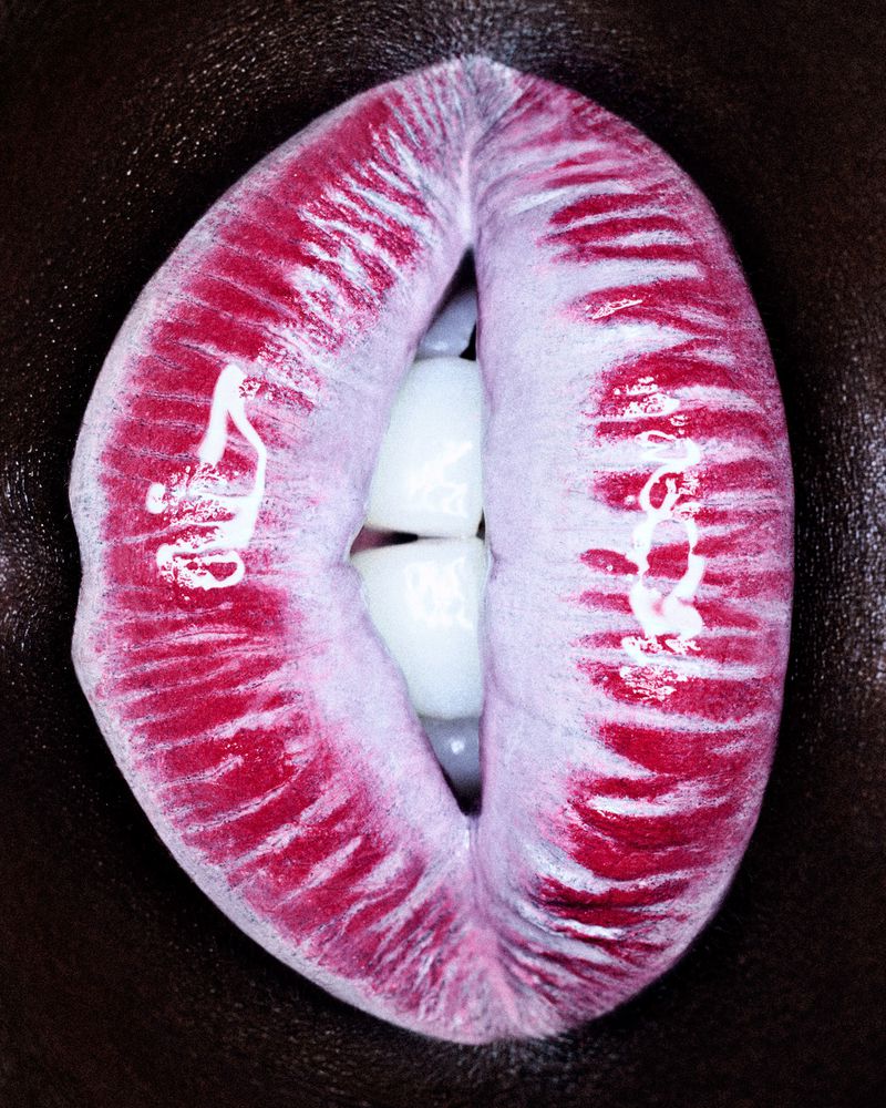 Close-up shot of a person's mouth featuring red lipstick and white teeth. The dark skin surrounding the mouth provides a contrasting backdrop.