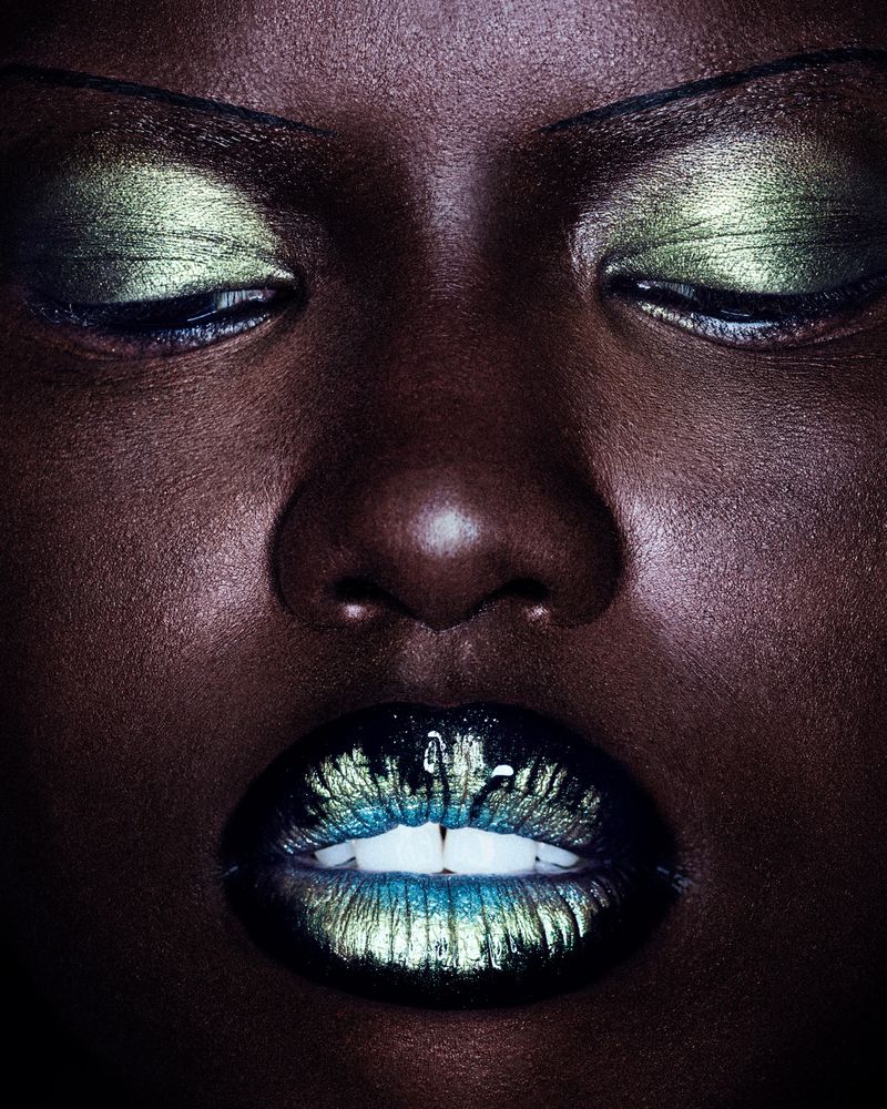 Close-up portrait of a woman with dark skin and closed eyes, vibrant green and blue eye shadow and matching lipstick, dark background.