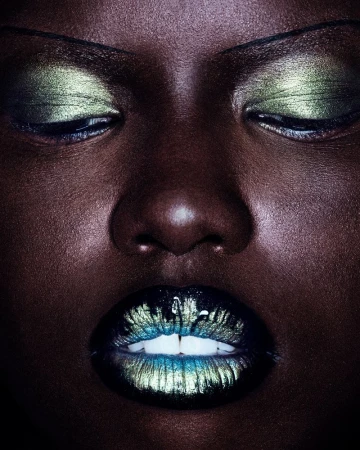 Close-up portrait of a woman with dark skin and closed eyes, vibrant green and blue eye shadow and matching lipstick, dark background.