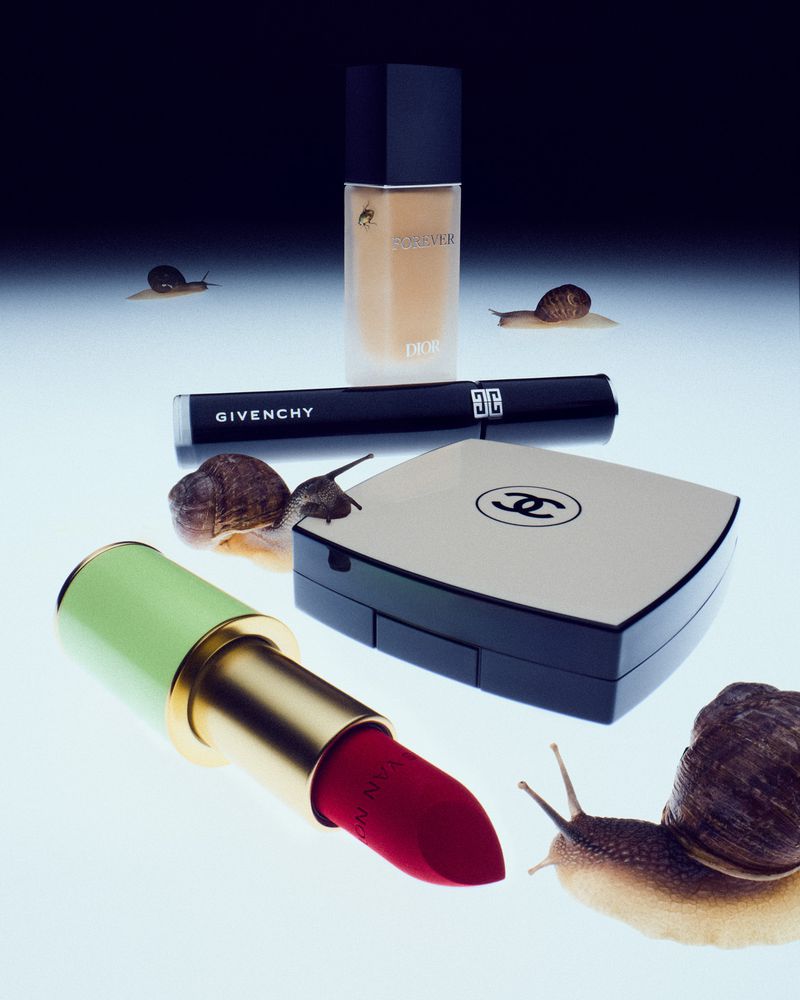 Still life shot of Dior foundation, Givenchy mascara, Chanel compact, and red lipstick, surrounded by live snails on a white surface.
