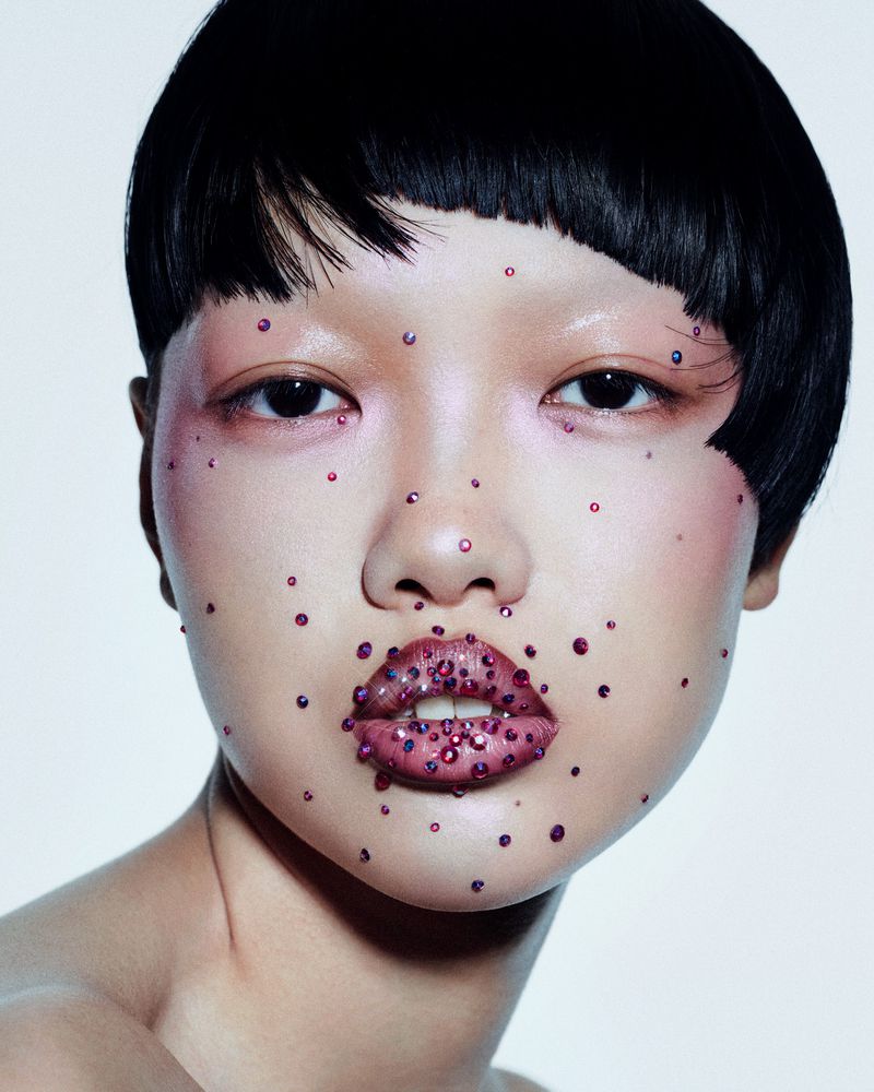 Close-up portrait of a young woman with short black hair, wearing decorative red gems on her face and lips against a neutral background.