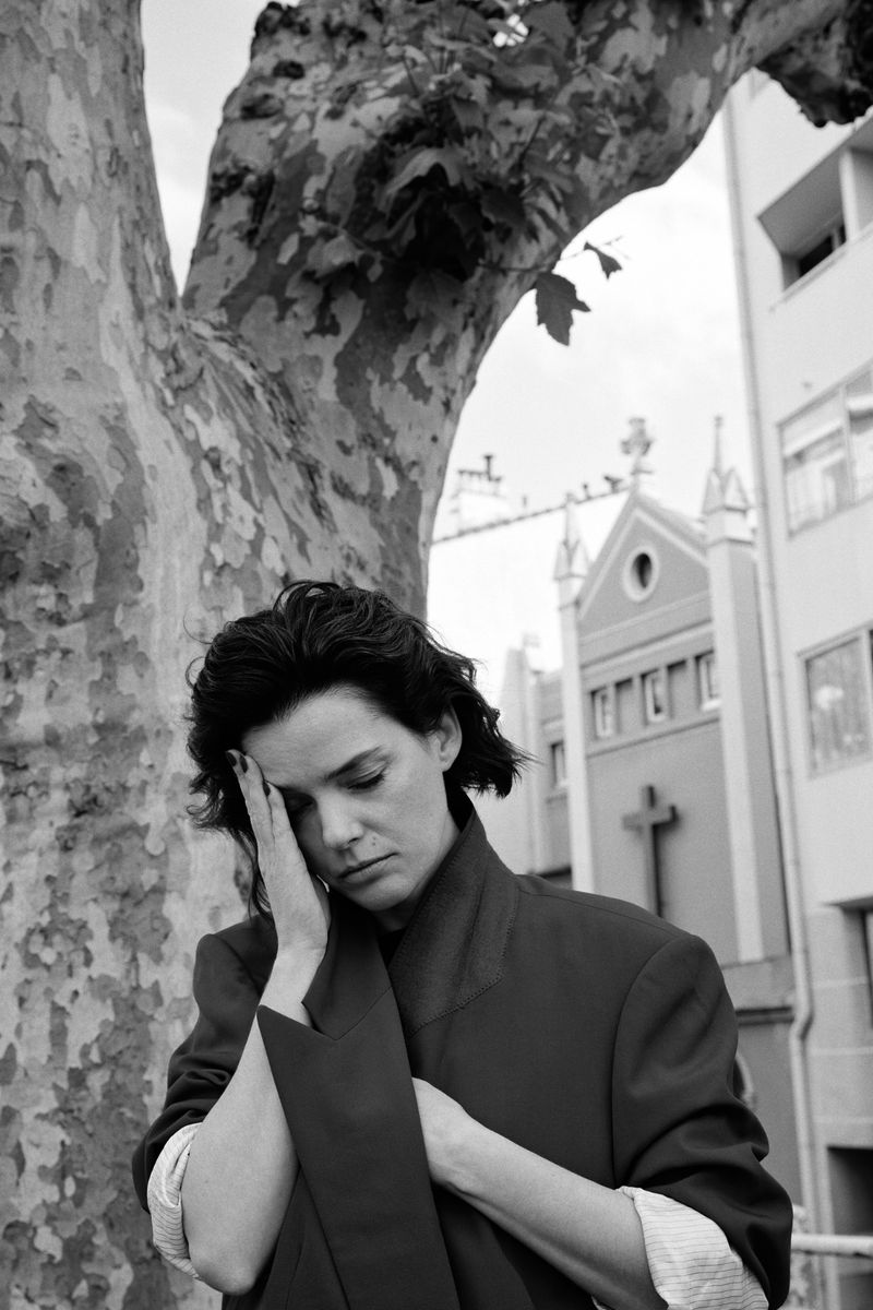 Black and white portrait of a woman with hands near her face, standing by a tree, with a church in the background on a cloudy day.