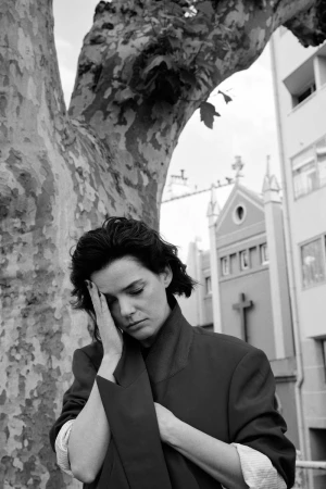 Black and white portrait of a woman with hands near her face, standing by a tree, with a church in the background on a cloudy day.