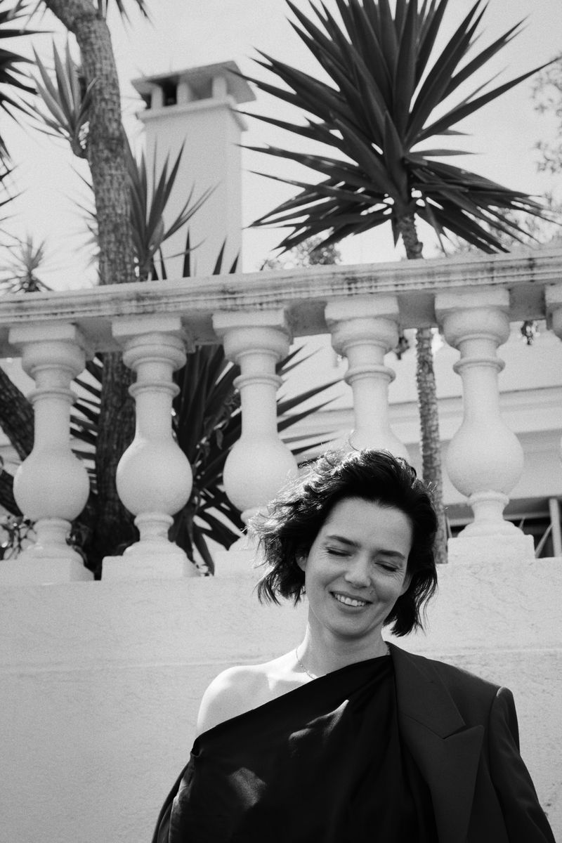 Black and white portrait of an adult woman with short hair smiling, wearing a jacket outdoors with palm trees in the background.