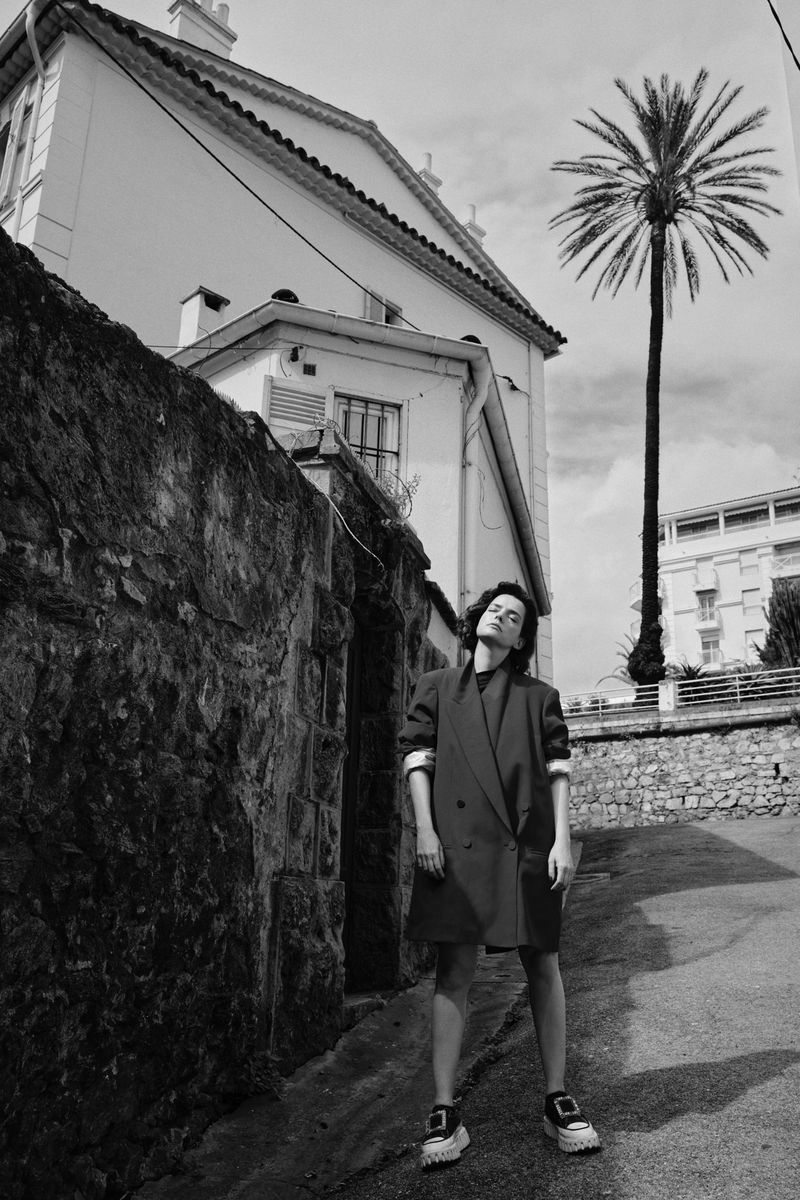 Black and white portrait of a woman in an oversized coat and sneakers standing in front of a stone wall and white building.