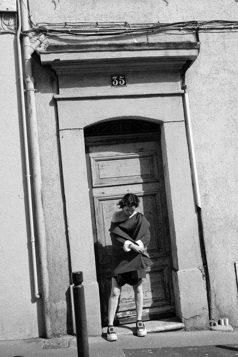 Monochrome portrait of a young adult woman standing in front of an old, weathered door on a building exterior in natural light.