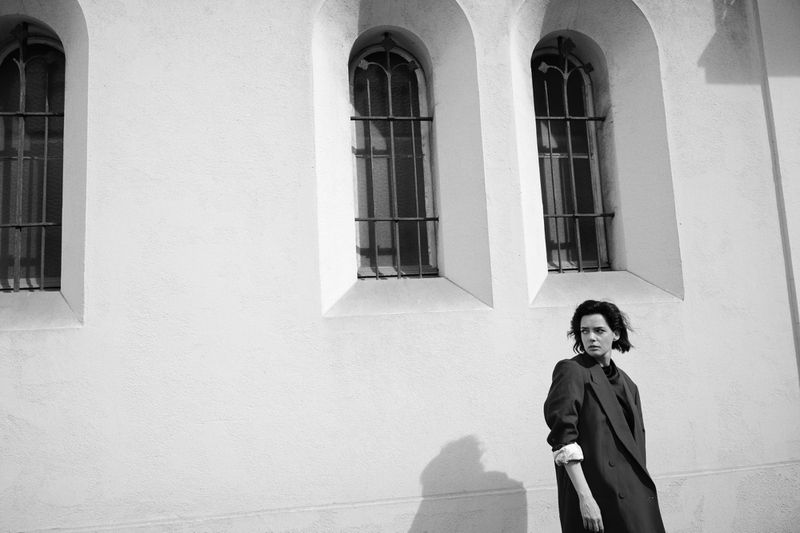 Monochrome outdoor portrait of a woman in a coat against a textured white wall with arched windows, creating a stark, fashionable image.