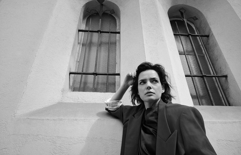 Monochrome portrait of a woman in a jacket, standing against a wall with barred windows looking up and to the left with a serious expression.